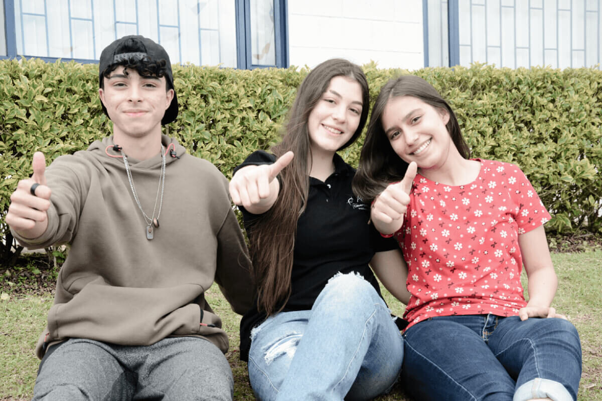 Fotografía de dos jóvenes alumnas y un alumno del Cibercolegio en el Campus de la Fundación Universitaria Católica del Norte, se encuentran sentados en la zona verde y reflejan sentirse felices disfrutando de su colegio. Una de las alumnas es la personera estudiantil electa en el año 2022.