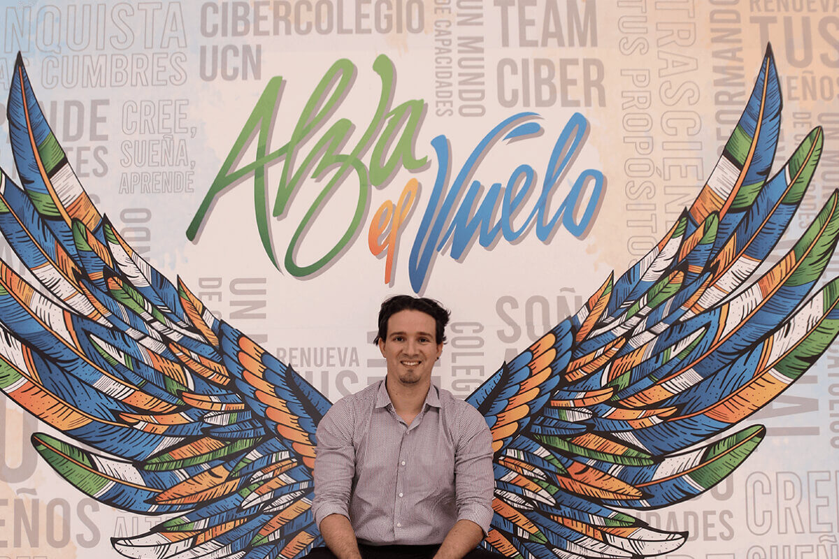 Joven sentado sonriendo y al fondo la imagen del logo del ciclo de la trascendencia que nos invita alzar el vuelo. El logo esta formado por dos alas de varios colores, abiertas y volando.