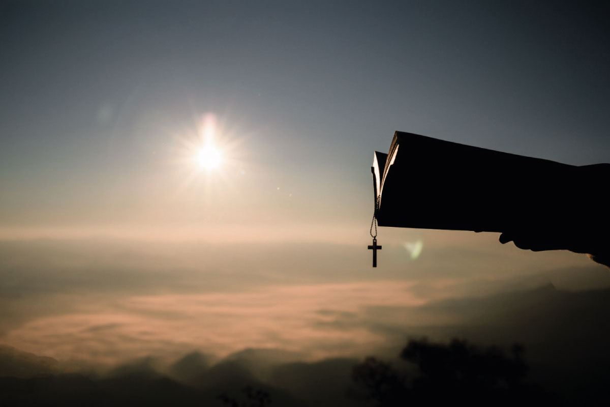 Imagen de un atardecer y una persona sosteniendo una Biblia y una cruz